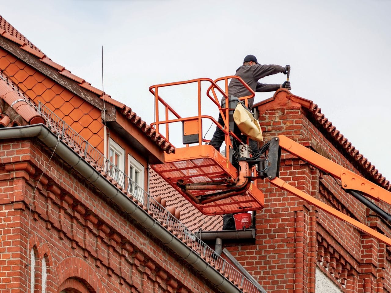 Un couvreur dans une nacelle intervient au bord d'un toit de tuiles, sur une façade en briques